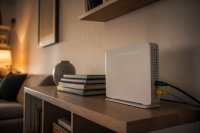 Heimrouter auf einem Wohnzimmerregal mit angeschlossenen Kabeln und leuchtenden Statusanzeigen - Auf einem hölzernen Wohnzimmerregal steht ein weißer Heimrouter mit zwei angesteckten Netzwerkkabeln und einer Reihe grün leuchtender Statusanzeigen, daneben ein Stapel Bücher, im Hintergrund eine unscharfe Wohnwand am Abend. Symbolbild zum Datenverkehr, der über private Internetanschlüsse läuft. Bild KI-generiert