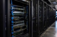 Serverschrank mit Netzwerkkabeln in einem abgedunkelten Technikraum - Ein geöffneter Serverschrank mit sortierten Netzwerkkabeln und Statusleuchten steht in einem abgedunkelten Technikraum, im Hintergrund verschwimmt eine Reihe weiterer Schränke. Symbolbild zum Wirtschaftsschutz in deutschen Unternehmen