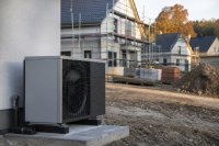 Neubausiedlung mit Einfamilienhäusern im Rohbau, vor einem Haus steht das Außengerät einer Luft-Wasser-Wärmepumpe auf einem Betonsockel - Eine Neubausiedlung am Ortsrand im Herbstlicht, mehrere Einfamilienhäuser im Rohbau mit Gerüsten, im Vordergrund ein frisch gesetztes Außengerät einer Luft-Wasser-Wärmepumpe auf einem Betonsockel neben der Hauswand, daneben Baumaterial und ein Erdhaufen. Symbolbild zum Wärmepumpenanteil im Wohnungsneubau 2025. Bild KI-generiert.