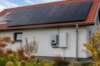 Solarmodule auf einem Ziegeldach mit Stromzähler und Wechselrichter an der Hauswand. Auf einem roten Ziegeldach eines Einfamilienhauses liegen dunkle Solarmodule, an der Hauswand darunter hängen ein Wechselrichter und ein Zählerkasten mit geschlossener Klappe, davor ein kleiner Garten im Herbstlicht. Symbolbild zur Einspeisevergütung und zum Eigenverbrauch von Solarstrom.