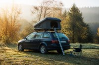 Kombi mit aufgeklapptem Hartschalen-Dachzelt auf einer Wiese am Waldrand, daneben ein Campingstuhl im Herbstlicht. Ein gewöhnlicher Kombi steht auf einer Wiese am Waldrand, auf dem Dach ist ein Hartschalen-Dachzelt aufgeklappt, eine Leiter lehnt an der Seite; daneben ein Campingstuhl und ein kleiner Gaskocher, das Laub im Hintergrund färbt sich herbstlich. Symbolbild zum Dachzelt als Alternative zum Reisemobil. Bild KI-generiert.