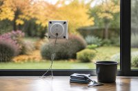 Fensterputzroboter arbeitet an einer großen Wohnzimmerscheibe, davor Eimer, Mikrofasertücher und ein Abzieher auf dem Boden. Ein quadratischer Fensterputzroboter haftet mit Sicherungsseil an einer bodentiefen Wohnzimmerscheibe, auf dem Parkett davor stehen ein Eimer, gefaltete Mikrofasertücher und ein Fensterabzieher, draußen unscharf ein herbstlicher Garten im Nachmittagslicht. Symbolbild zu Haushaltshilfen und Haushaltsgeräten im Oktober 2026.