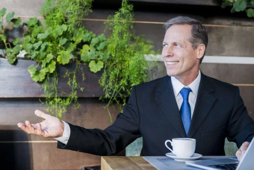 smiling-mature-businessman-making-hand-gesture-with-cup-coffee-laptop-desk-scaled-1-2048x1367.j.webp