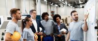Mehrere Personen stehen in einem industriellen Arbeitsumfeld gemeinsam vor einem Board und diskutieren Verbesserungsthemen im Rahmen des KVP. Auf dem Board sind Prozesse, Notizen und visuelle Darstellungen zur Analyse von Abläufen zu erkennen. Die Szene vermittelt einen strukturierten Austausch zur kontinuierlichen Verbesserung von Arbeitsprozessen. Im Mittelpunkt steht die gemeinsame Bewertung von Herausforderungen und die Entwicklung praxisnaher Lösungen im Team.