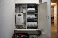 Ein geöffneter Zählerschrank in einem Hausanschlussraum, darin ein elektronischer Stromzähler mit Display, ein kleines Kommunikationsmodul mit Leuchtdioden und Reihen beschrifteter Sicherungsautomaten, daneben ein Werkzeugkoffer auf dem Boden, nüchternes Kellerlicht. Symbolbild zum Smart-Meter-Rollout im Oktober 2026. Bild KI-generiert.